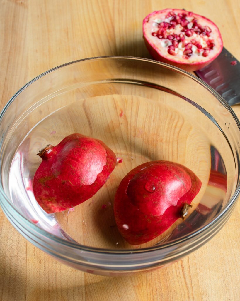 A pomegrate sliced in half and chunks of pomegranate floating in a glass bowl of water.