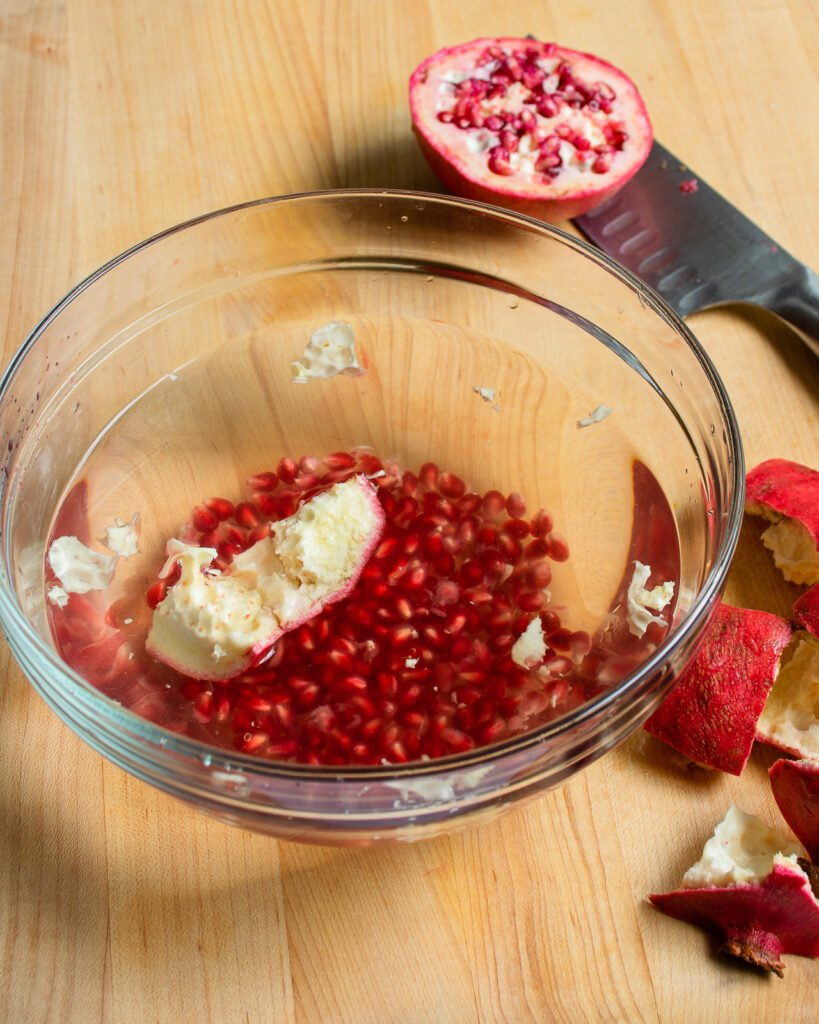 Pomegranate seeds in a glass bowl of water and a chunk of pomegranate floating on top.