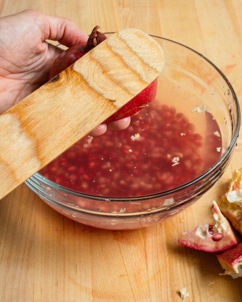 A hand banging the back of a halved pomegranate with a wooden spoon.