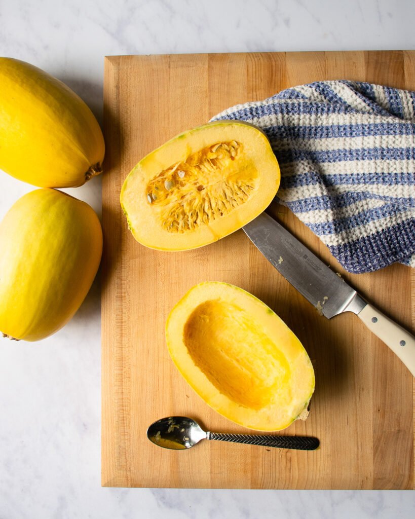Spaghetti squash on a cutting board with a white handled knife, a blue and white striped towel, a spoon.