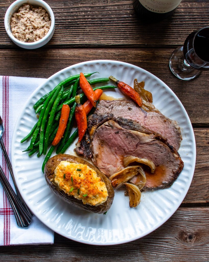 A dinner plate with roast beef, a twice baked potato and some vegetables on a wooden table.