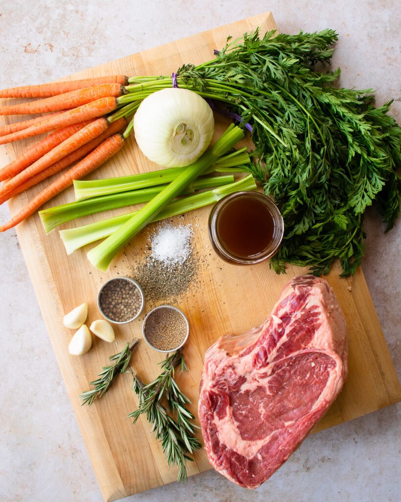 Ingredients on a cutting board - carrots, celery, onion, herbs, stock, seasonings and a beef rib roast.