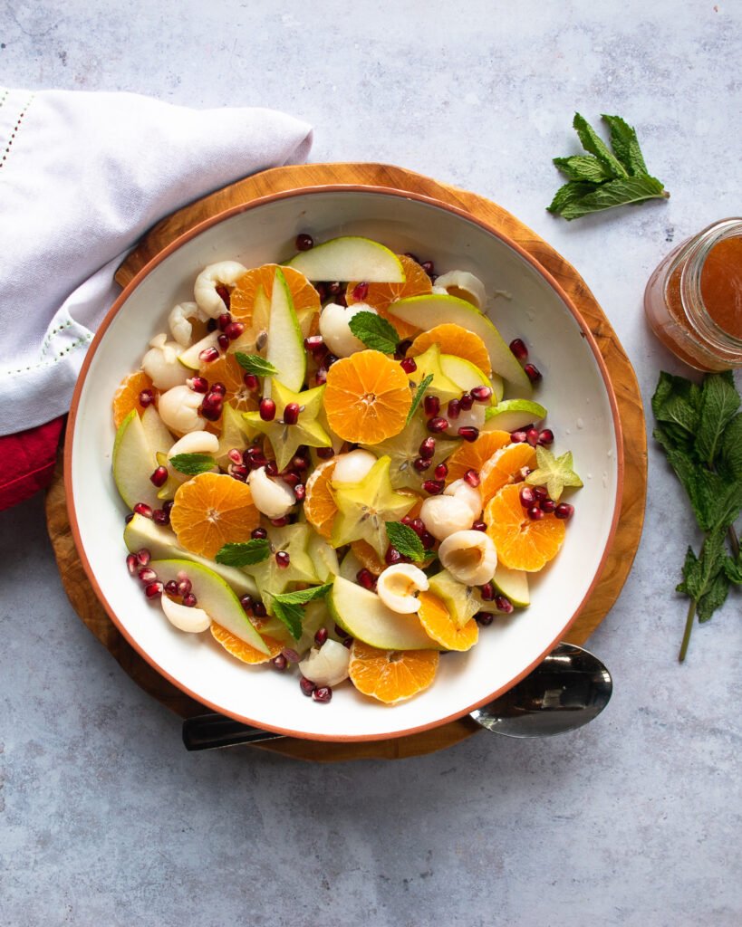Winter Fruit Salad in a white bowl on a wooden charger with mint leaves and a jar of dressing near by.