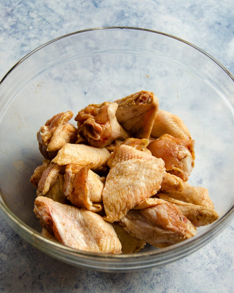 Chicken wings, tossed with oil and Chinese 5-Spice Powder in a glass bowl on a blue marble counter.