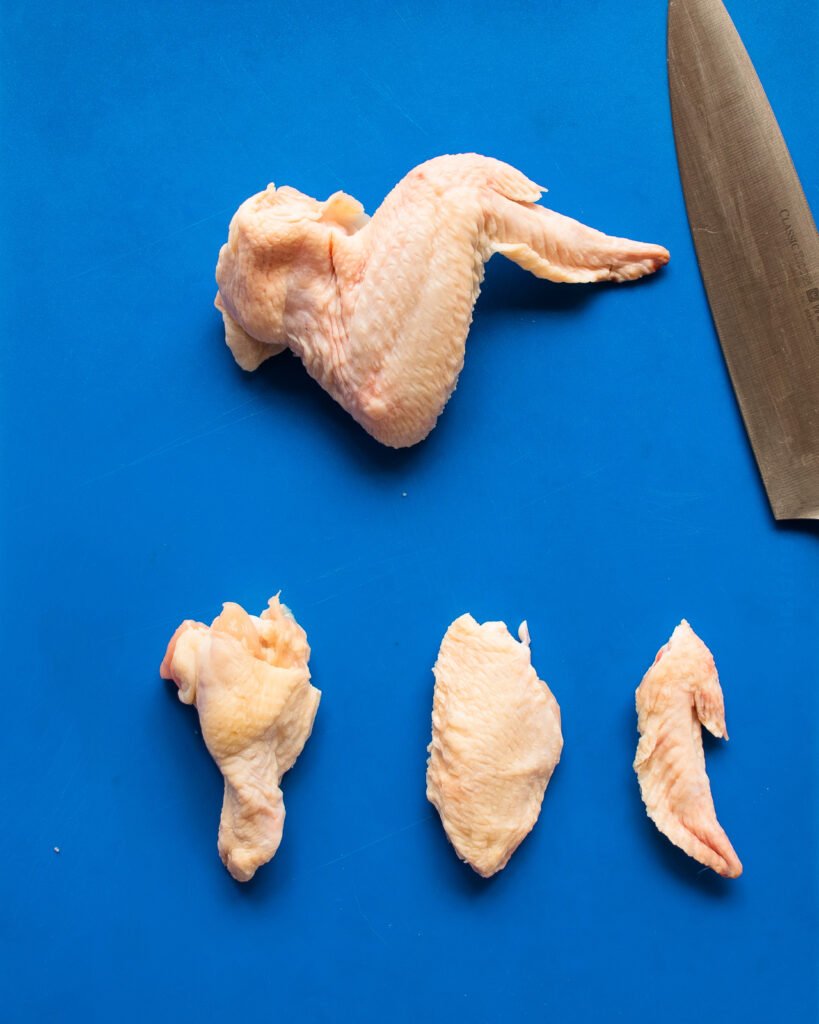 A chicken wing on a blue cutting board with another chicken wing cut into segments below it, along  with a white-handled knife.