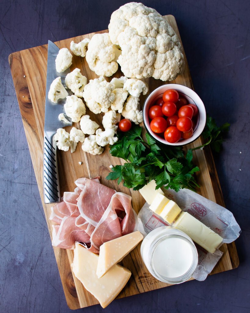 Ingredients on a cutting board - cauliflower, prosciutto, tomatoes, parsley, cream, butter and Parmesan cheese.
