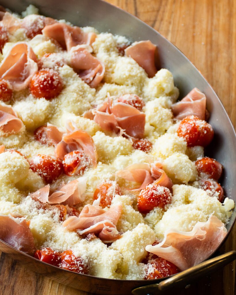Close up of cauliflower Alfredo bake before being sent to the oven - prosciutto and roasted tomatoes tucked into cauliflower with cheese sprinkled on top.