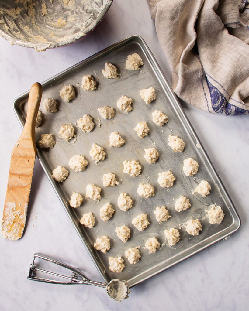 Small spoonfuls of biscuit dough on a baking sheet with a wooden spoon and towel near by.