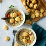 Two bowls of slow cooker chicken pot pie soup with more biscuit croutons on a wooden board and a blue napkin next to it.