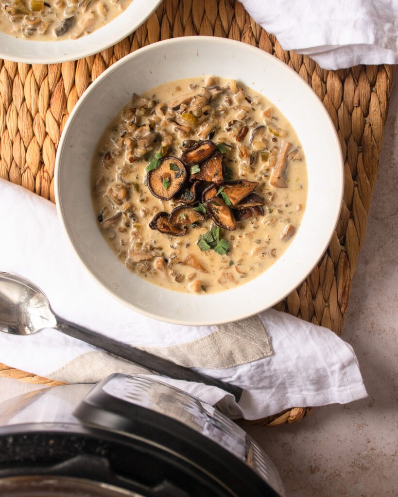 A bowl of cream of mushroom soup on a rattan mat with napkin and spoon and a pressure cooker.