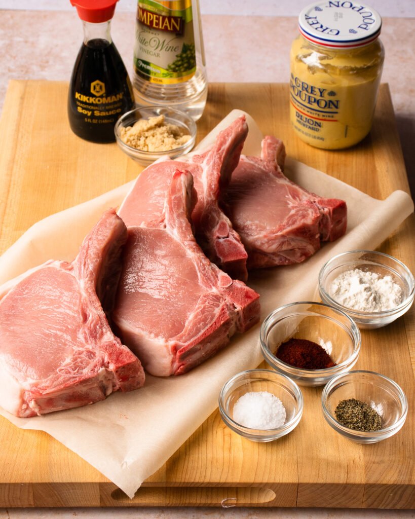 Ingredients for brown sugar pork chops on a wooden cutting board - flour, paprika, salt and pepper, mustard, vinegar, soy sauce and brown sugar.