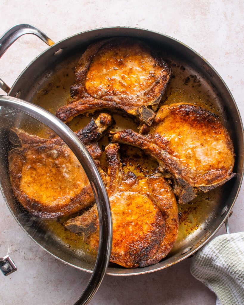 Four seasoned pork chops in a saute pan with a lid askew.