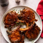 Four skillet glazed pork chops on a white plate with a red napkin beside it.