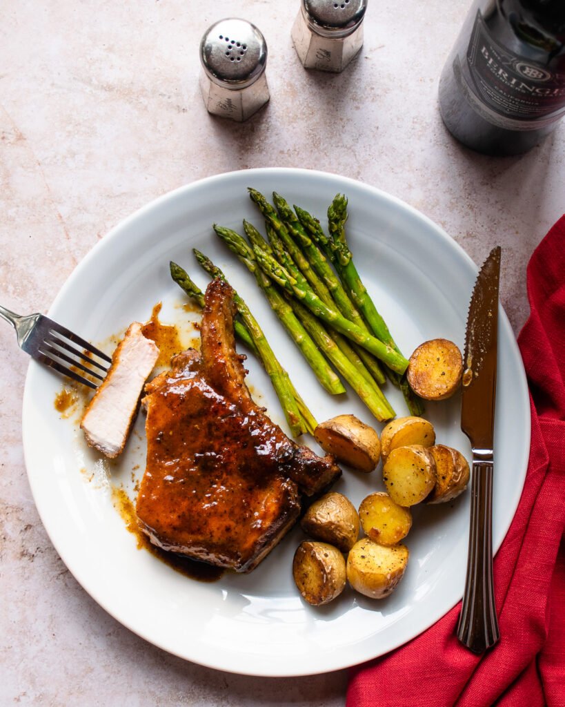 A white dinner plate with a pork chop, some potatoes and asparagus, along with a bottle of wine, salt and pepper shakers and a red napkin.