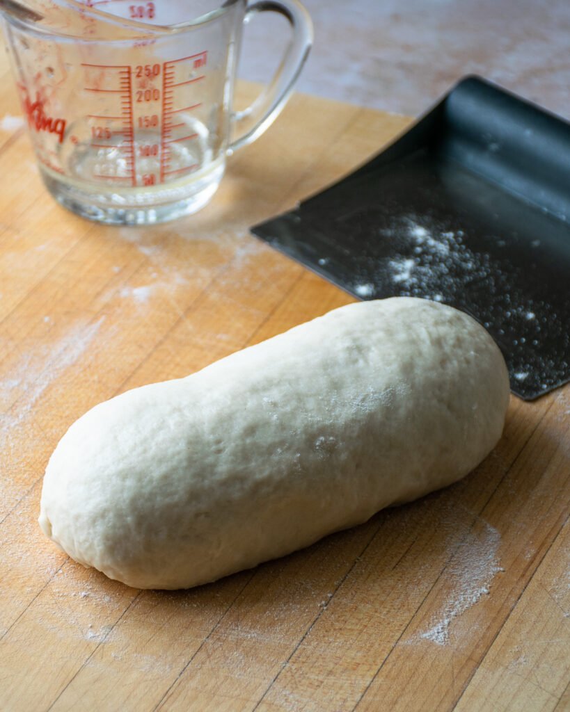 A log of dough on a cutting board with a bench scraper and a measuring glass.