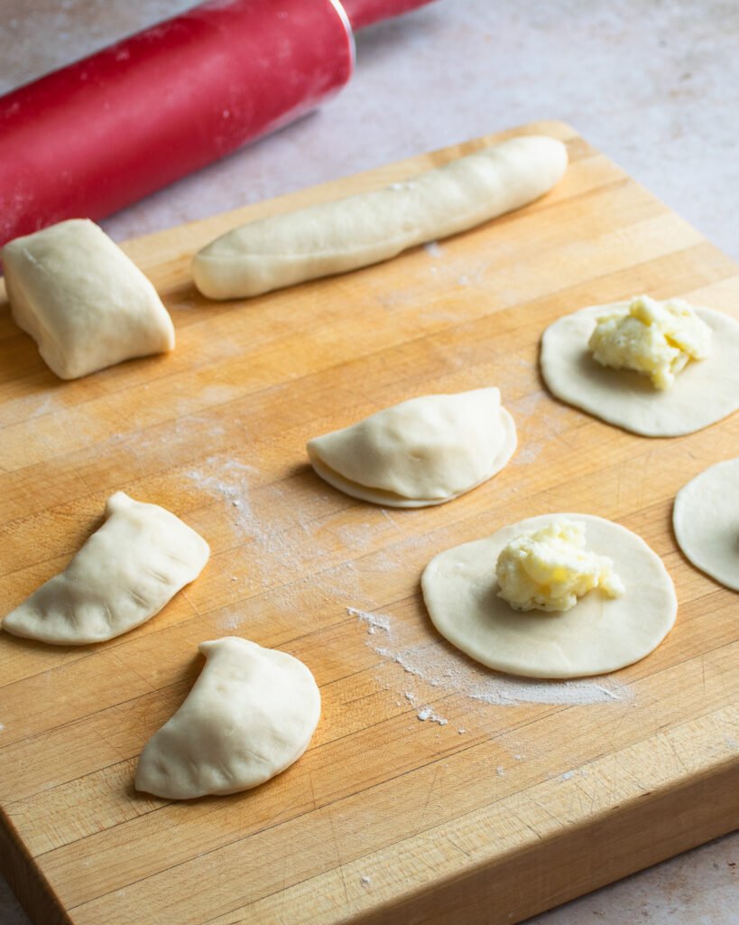 Two pierogies finished on a cutting board with four more doughs being made.