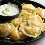 A dark blue plate with pierogies on it and a bowl of sour cream in the middle.