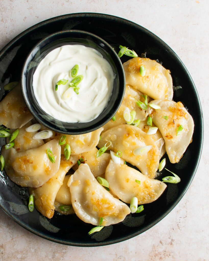 A dark blue plate of pierogies with scallions and a bowl of sour cream.