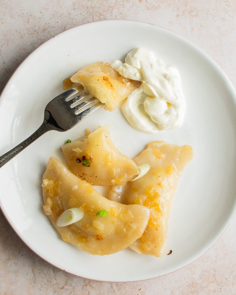 A white plate with pierogies and sour cream and a fork.