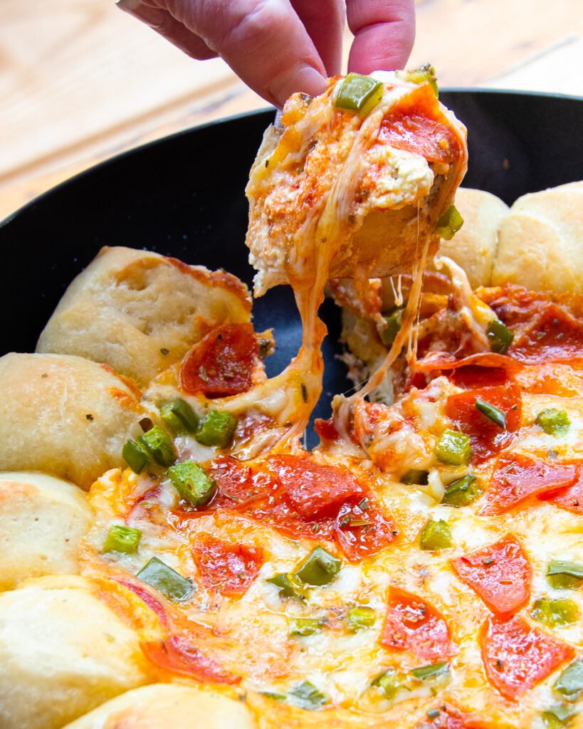 A dough ball with pizza dip being lifted out of a skillet with fingers.