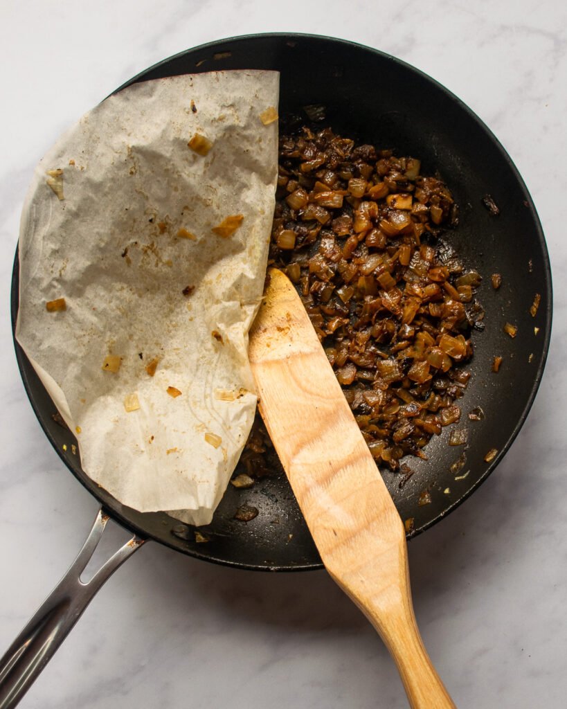 Caramelized onions in a skillet with a parchment circle peeled away and a wooden spoon inside.