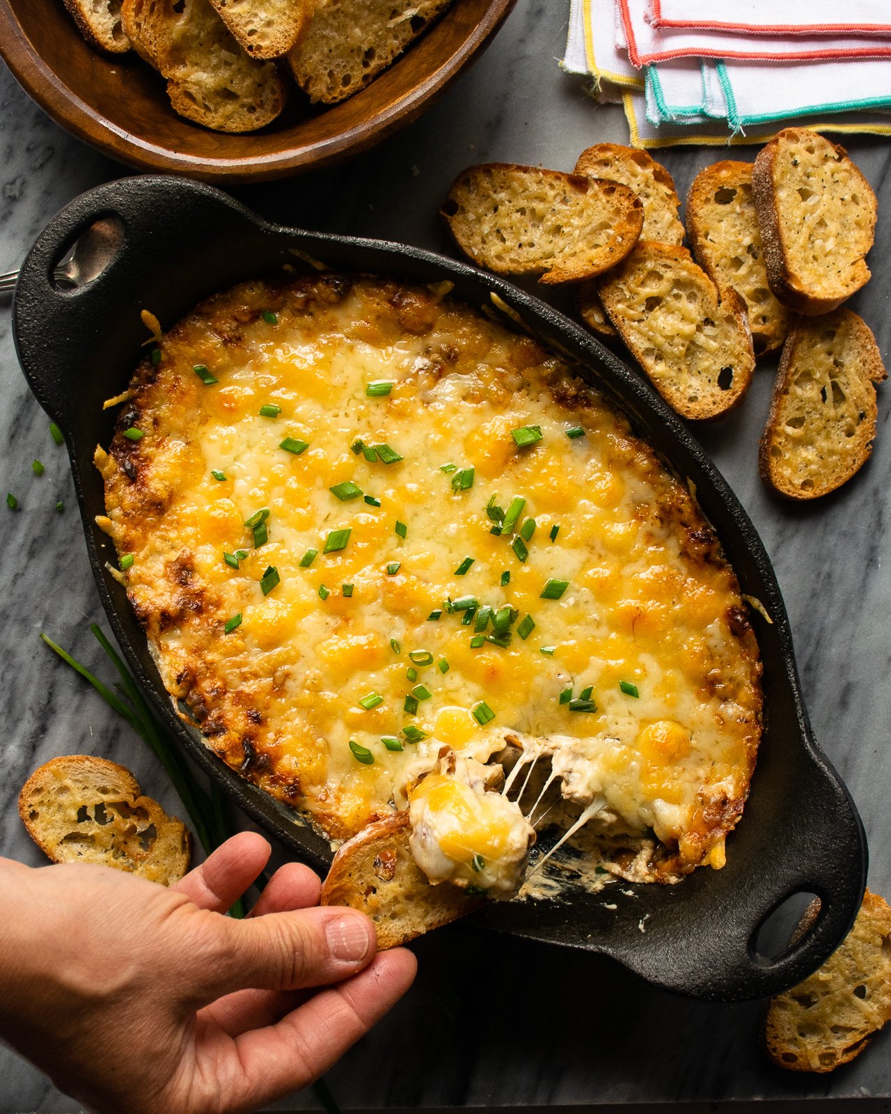 A hand dipping a crostini into warm french onion dip, with more crostini near by.