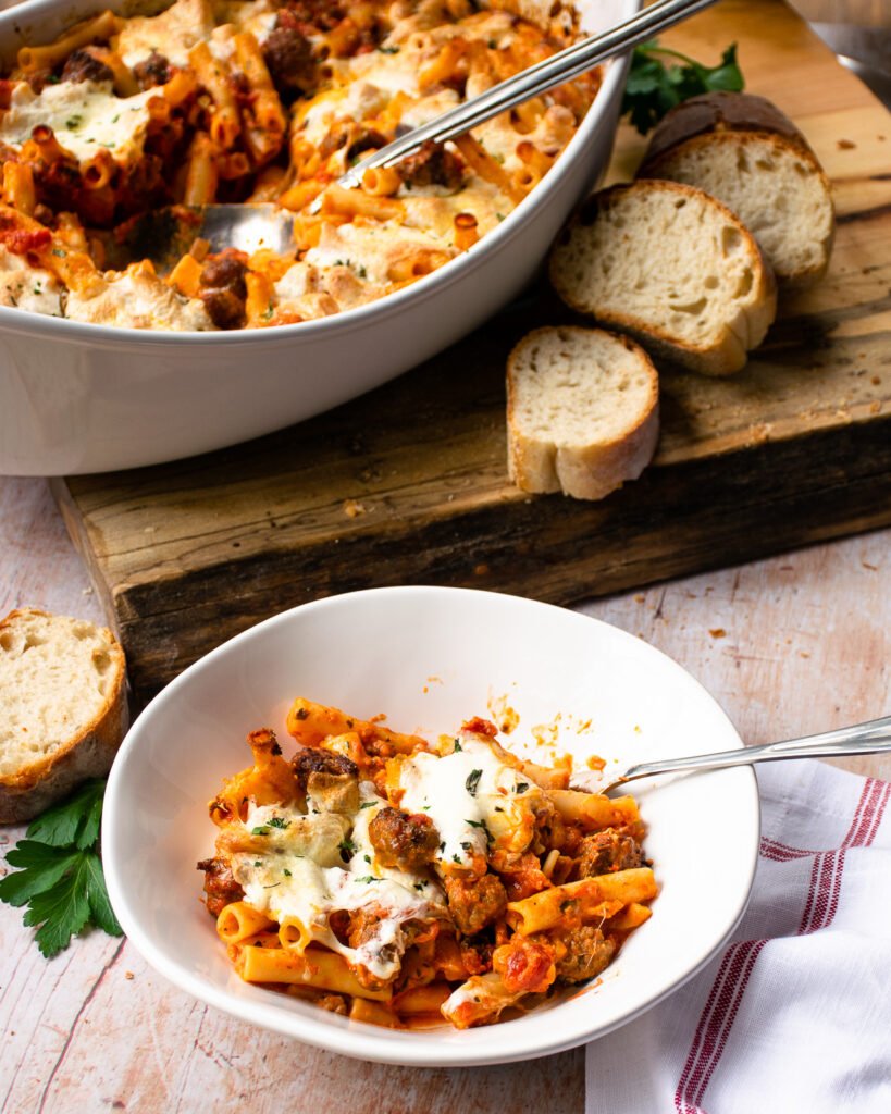 A bowl of ziti al forno next to a large white ceramic dish with more ziti al forno inside and a few slices of bread.