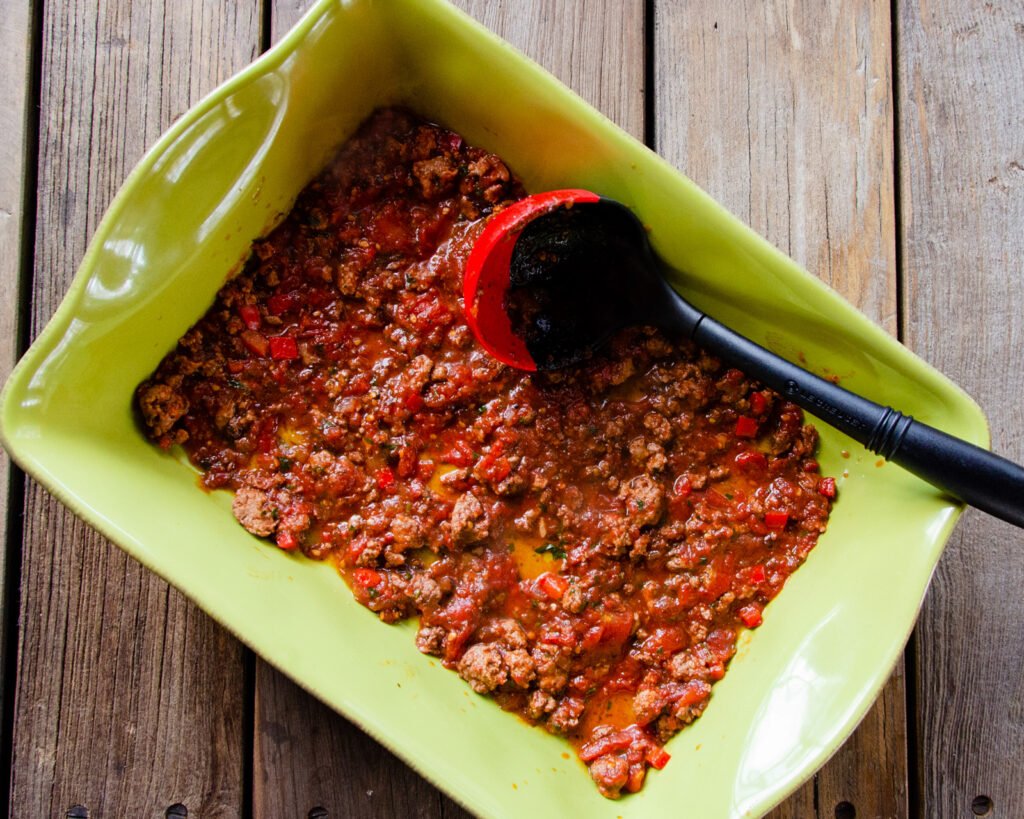 Tomato meat sauce in the bottom of a green ceramic casserole dish.