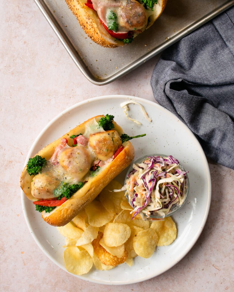 A white plate with an Italian chicken meatball grinder, some potato chips and some coleslaw next to a baking sheet with more sandwiches.