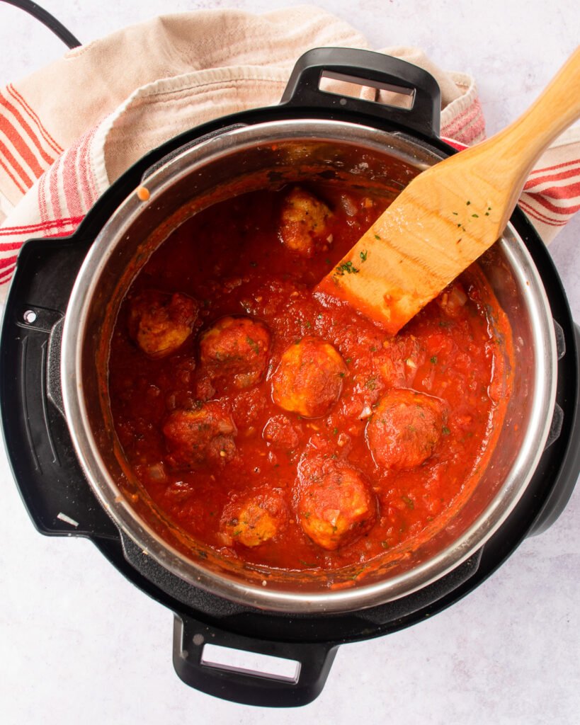 Meatballs and marinara in a pressure cooker with a wooden spoon sticking out of it.
