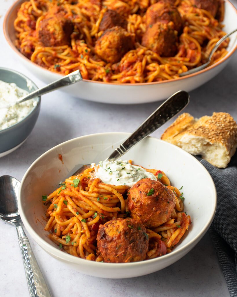 A white bowl of pressure cooker spaghetti and meatballs with whipped ricotta dolloped on top and a large bowl of more spaghetti in the background.