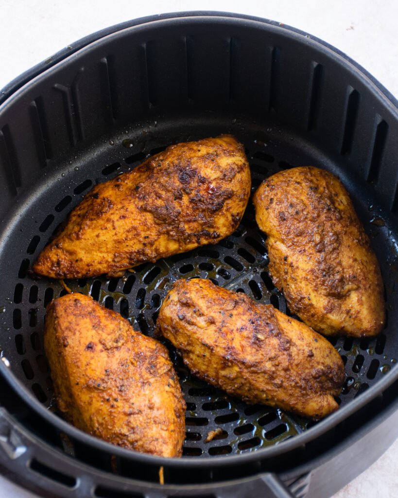 Four spiced chicken breasts in the basket of an air fryer.