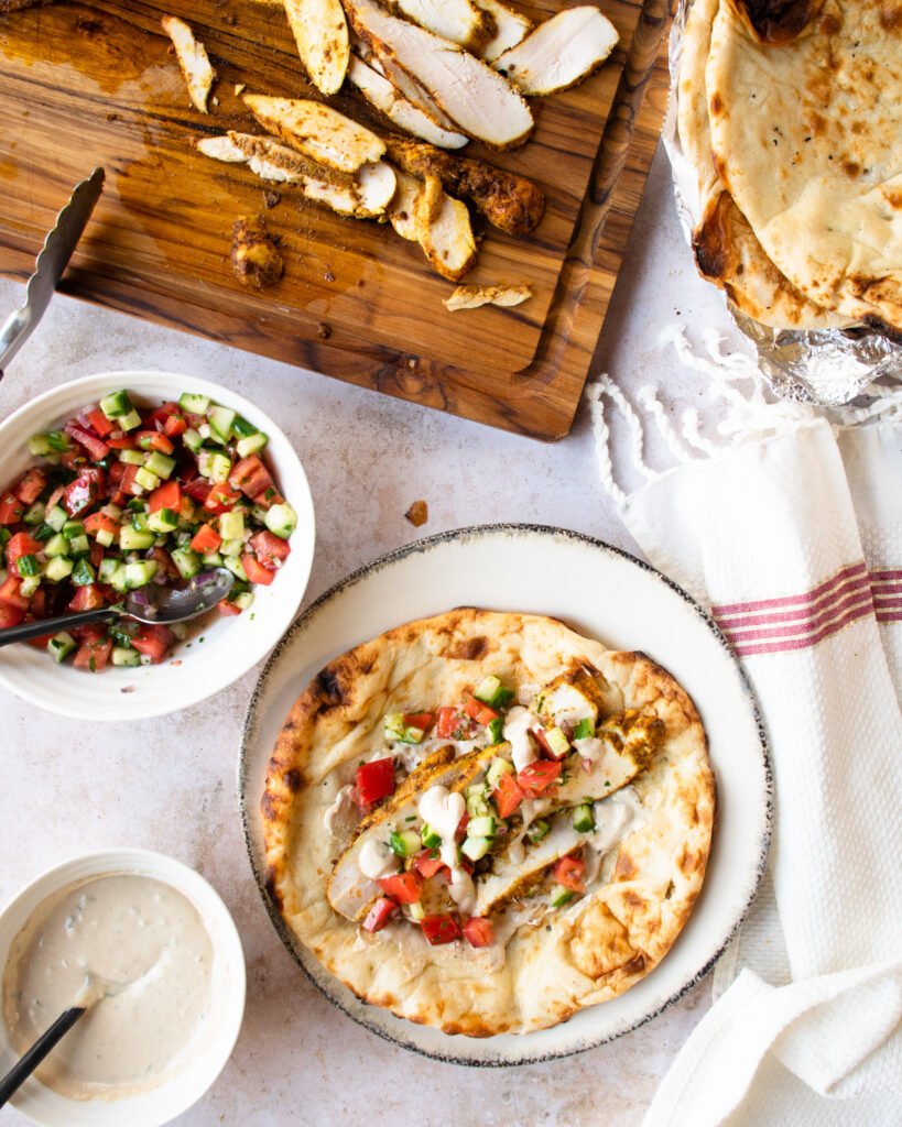 All the ingredients for chicken shawarma - naan, tomato cucumber salad, tahini sauce and sliced chicken on a cutting board.