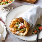 A chicken shawarma sandwich, half-wrapped in parchment paper on a white plate with ingredients around it.
