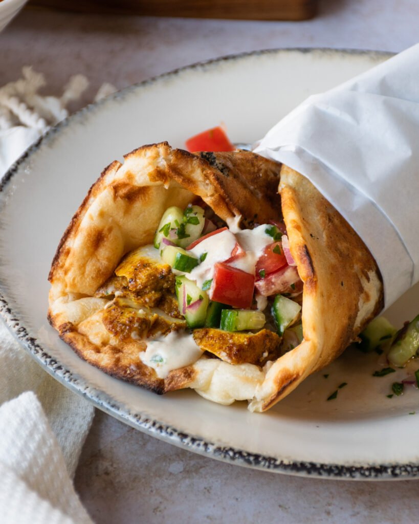 Close up shot of a chicken shawarma sandwich, half wrapped in parchment paper on a plate.