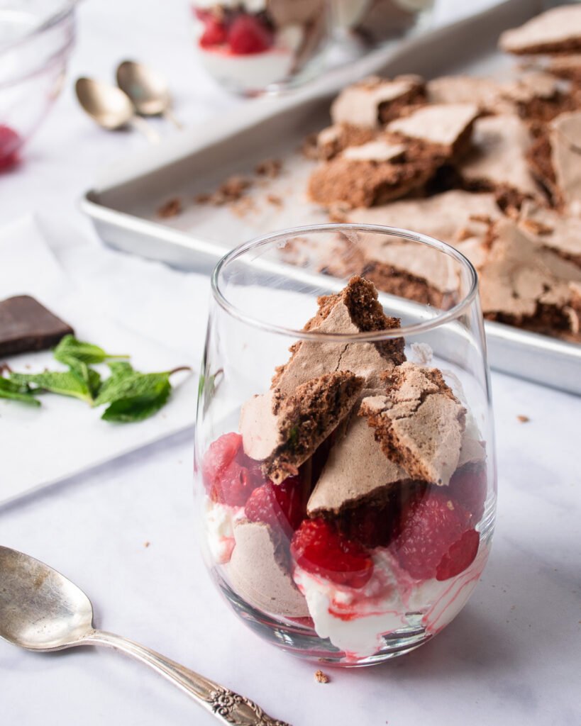 Chocolate Eton Mess in a stemless wine glass with broken meringue in the background.
