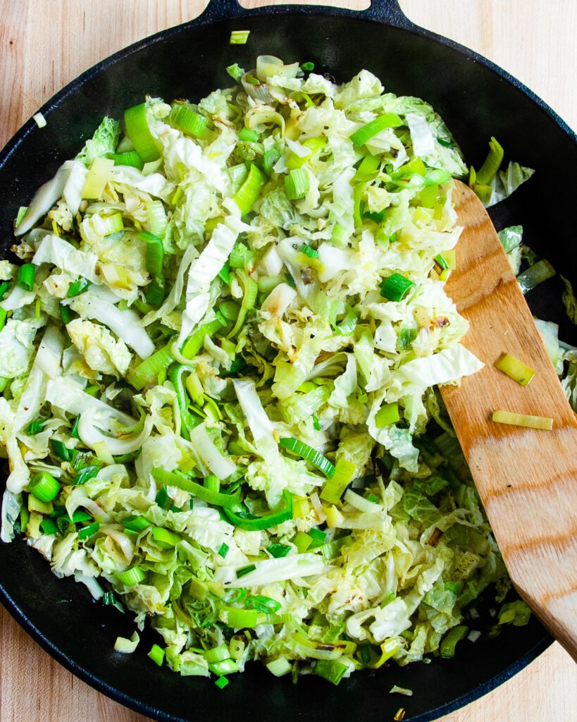 Leeks and cabbage wilting in a large cast iron skillet with a wooden spoon.
