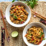 Two bowls of chicken drunken noodles on a placemat with chopsticks, half a lime and fresh Thai basil.