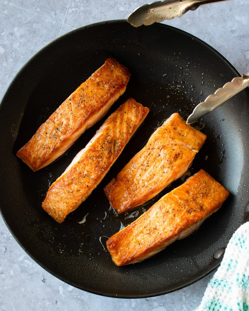 Four fillets of salmon in a non stick skillet with a pair of tongs and a hand towel.