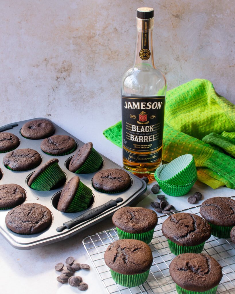 Chocolate cupcakes on the counter and in a muffin pan with a bottle of Irish whiskey and a green towel in the backgrouind.