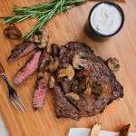 Marinated Rib Eye Steak on a cutting board with horseradish cream in a small dish and steak fries on the side.