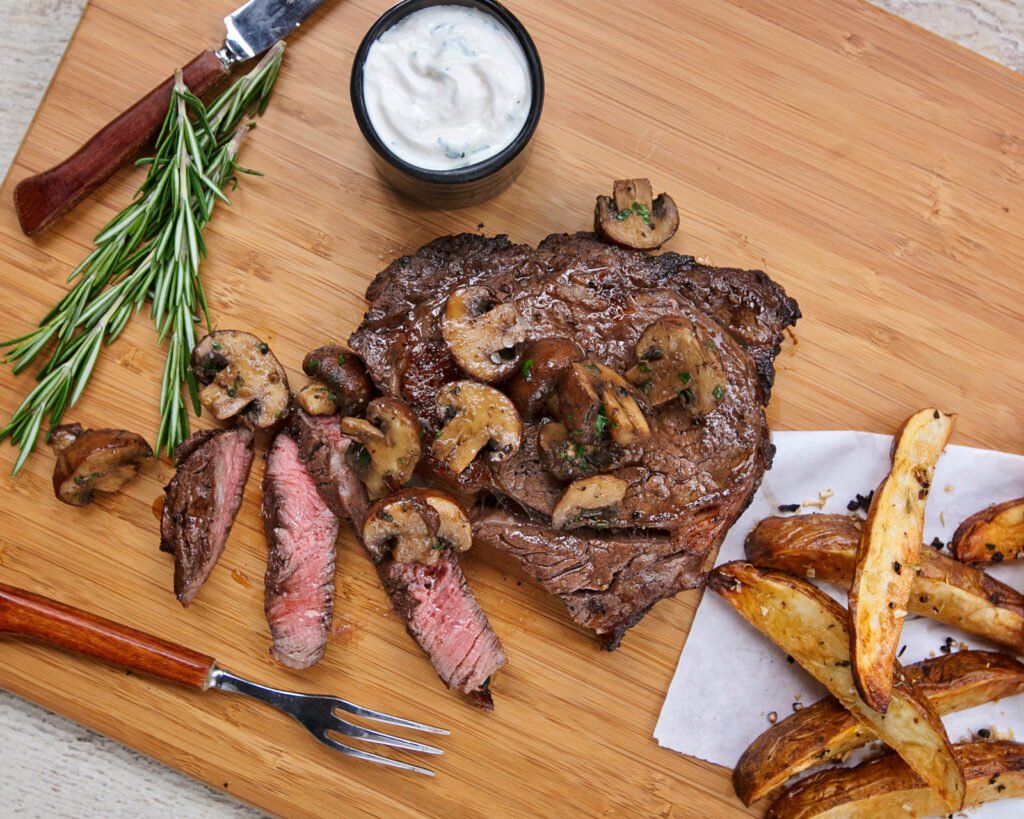 Marinated rib eye steak on a cutting board with knife and fork, horseradish cream and steak fries on the side.