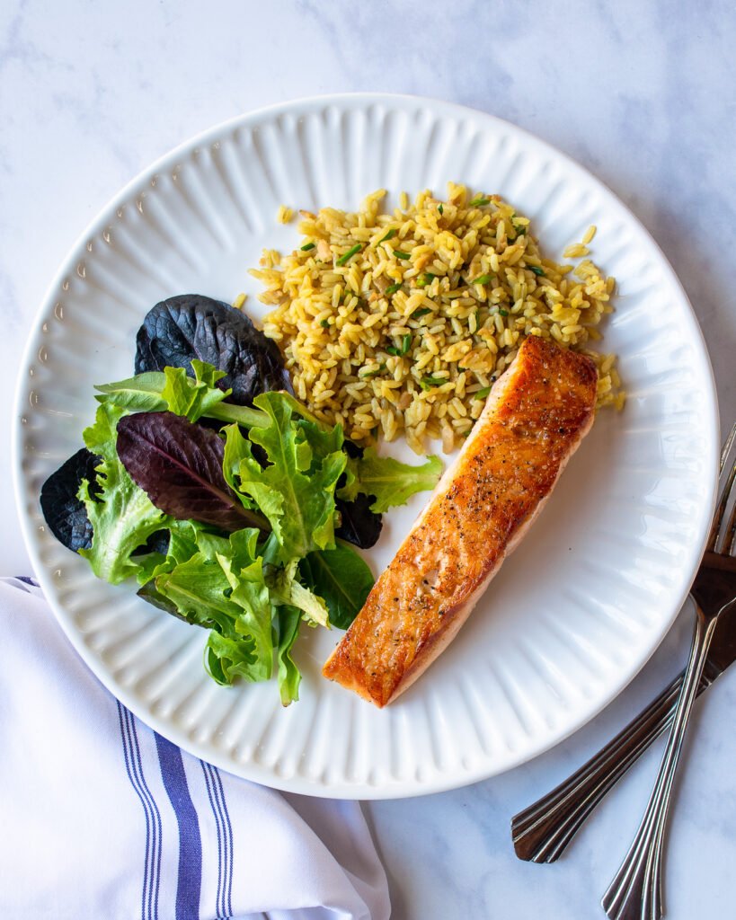 A fillet of pan roasted salmon on a white dinner plate with rice and salad greens.