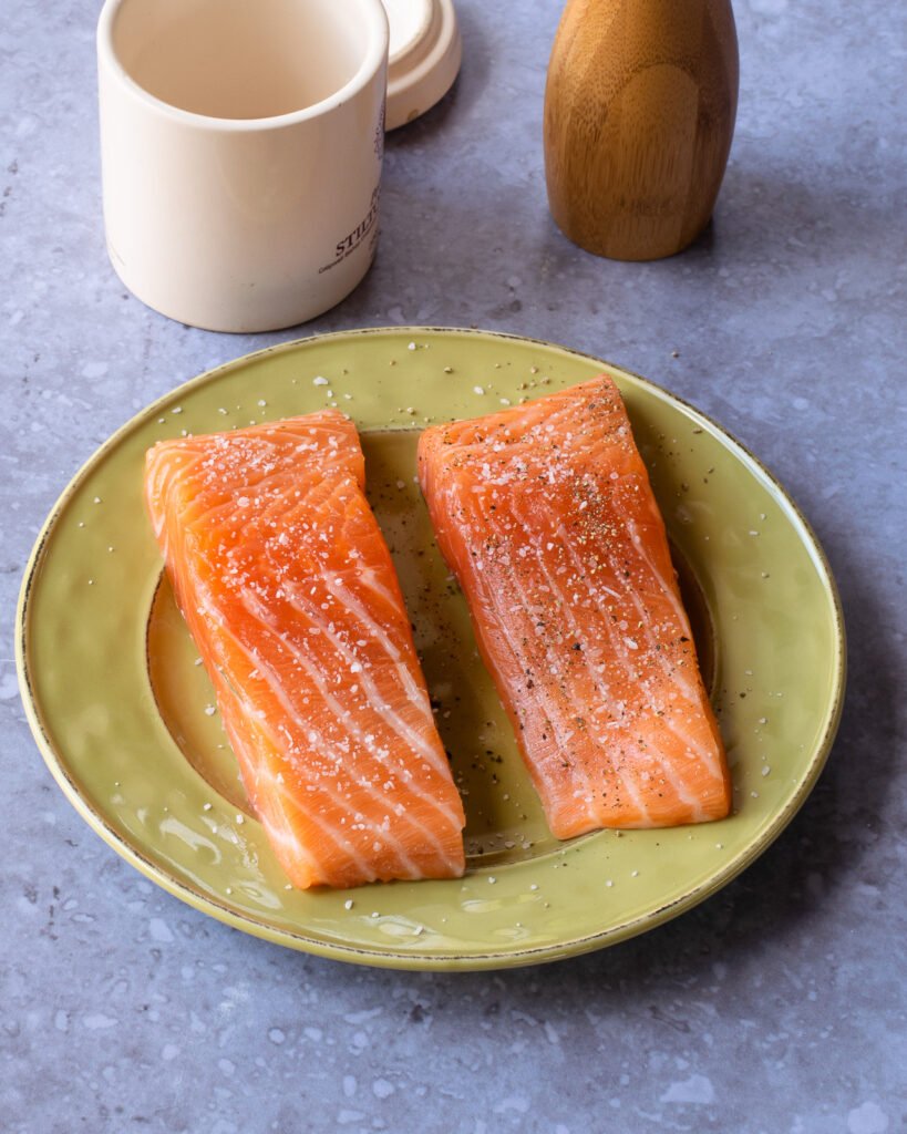 Two fillets of salmon on a green plate with salt and pepper in the background.