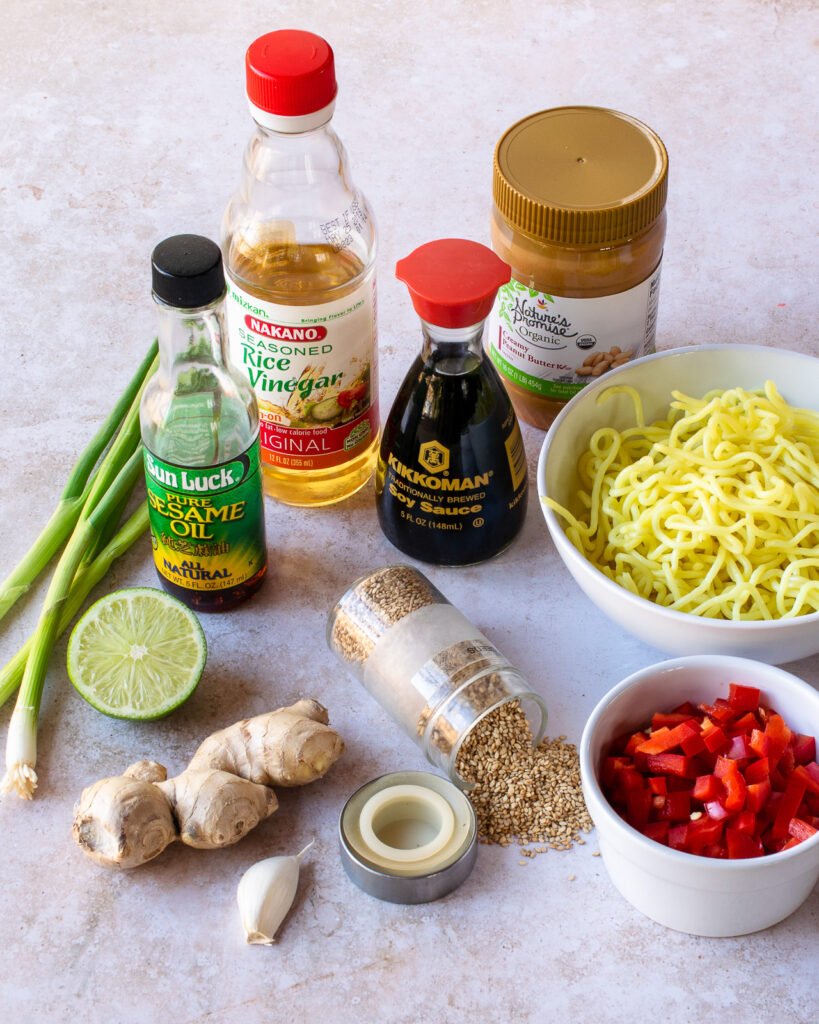 Ingredients on a counter - ginger, lime, garlic, sesame seeds, scallions, red pepper, noodles, rice wine vinegar, sesame oil, soy sauce and peanut butter.