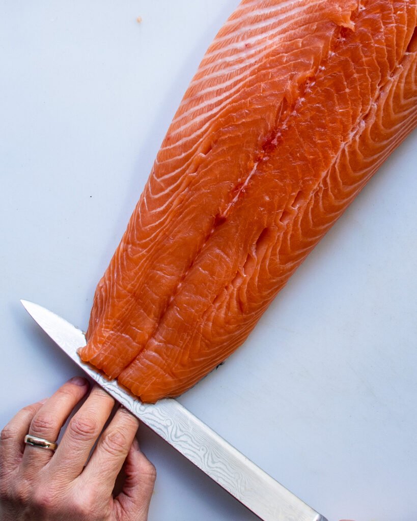 A carving knife starting to cut the tail end of a side of salmon on a white board.