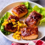 Four fillets of salmon with BBQ sauce on them, on a white plate with mango salsa and some lettuce leaves.