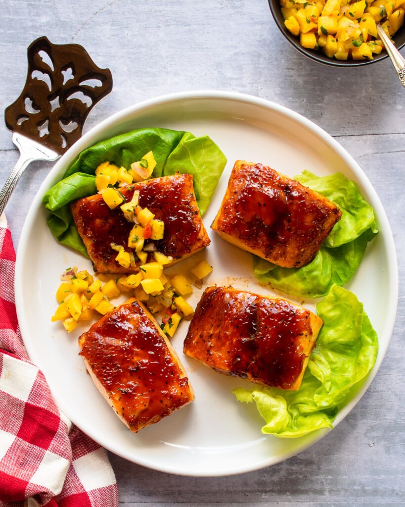 Balsamic BBQ Salmon on a white plate with lettuce and mango salsa, a spatula and red checkered napkin near by.