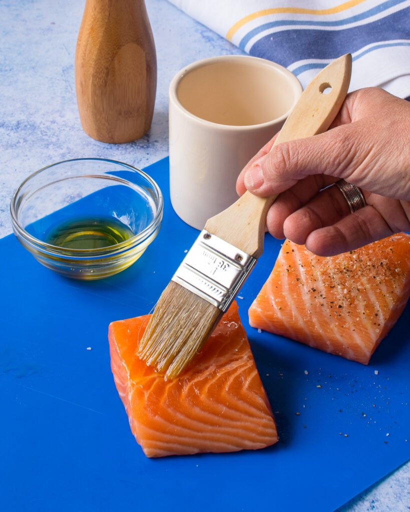 A hand brushing oil onto two fillets of salmon on a blue cutting board.