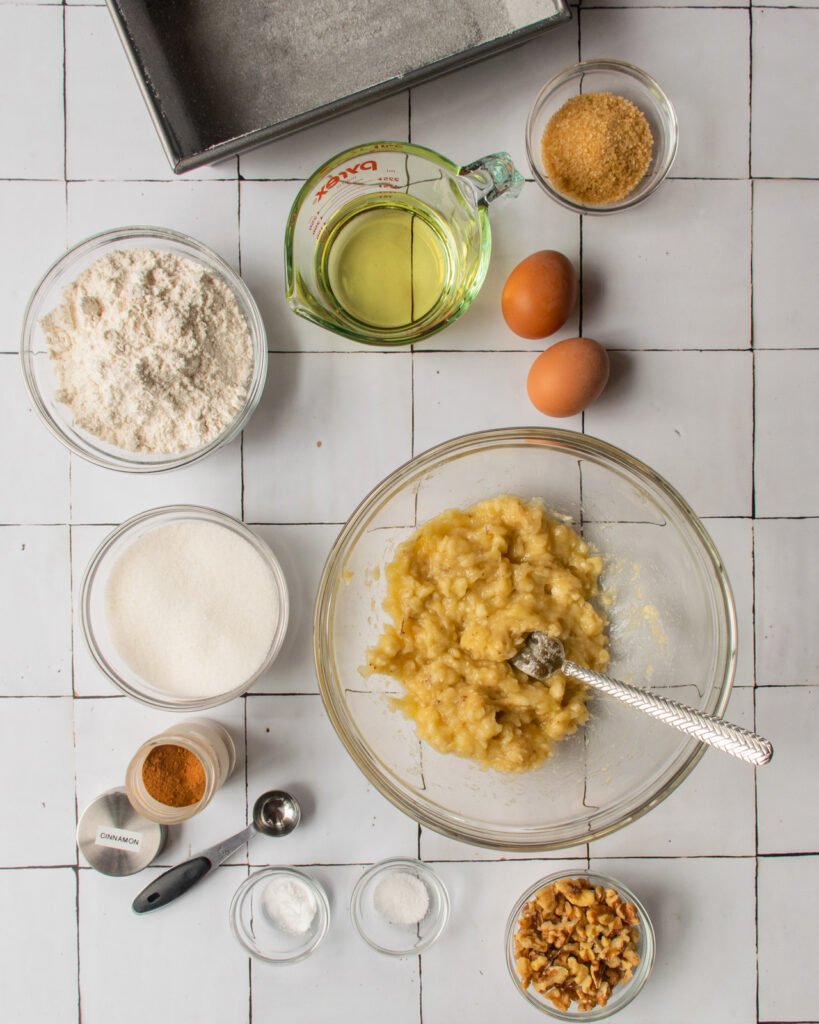 Ingredients on a counter - mashed banana, walnuts, baking soda, salt, cinnamon, salt, flour, vegetable oil and eggs.